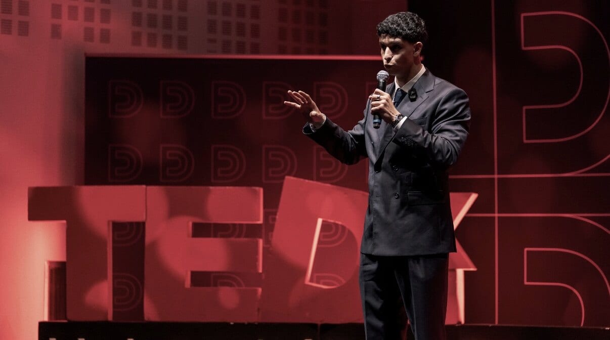 Naïm Khelif au micro sur la scène du TEDx Paris Dauphine, devant les lettres TEDx.